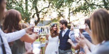 Guests with smartphones taking photo of bride and groom at wedding reception outside in the backyard.