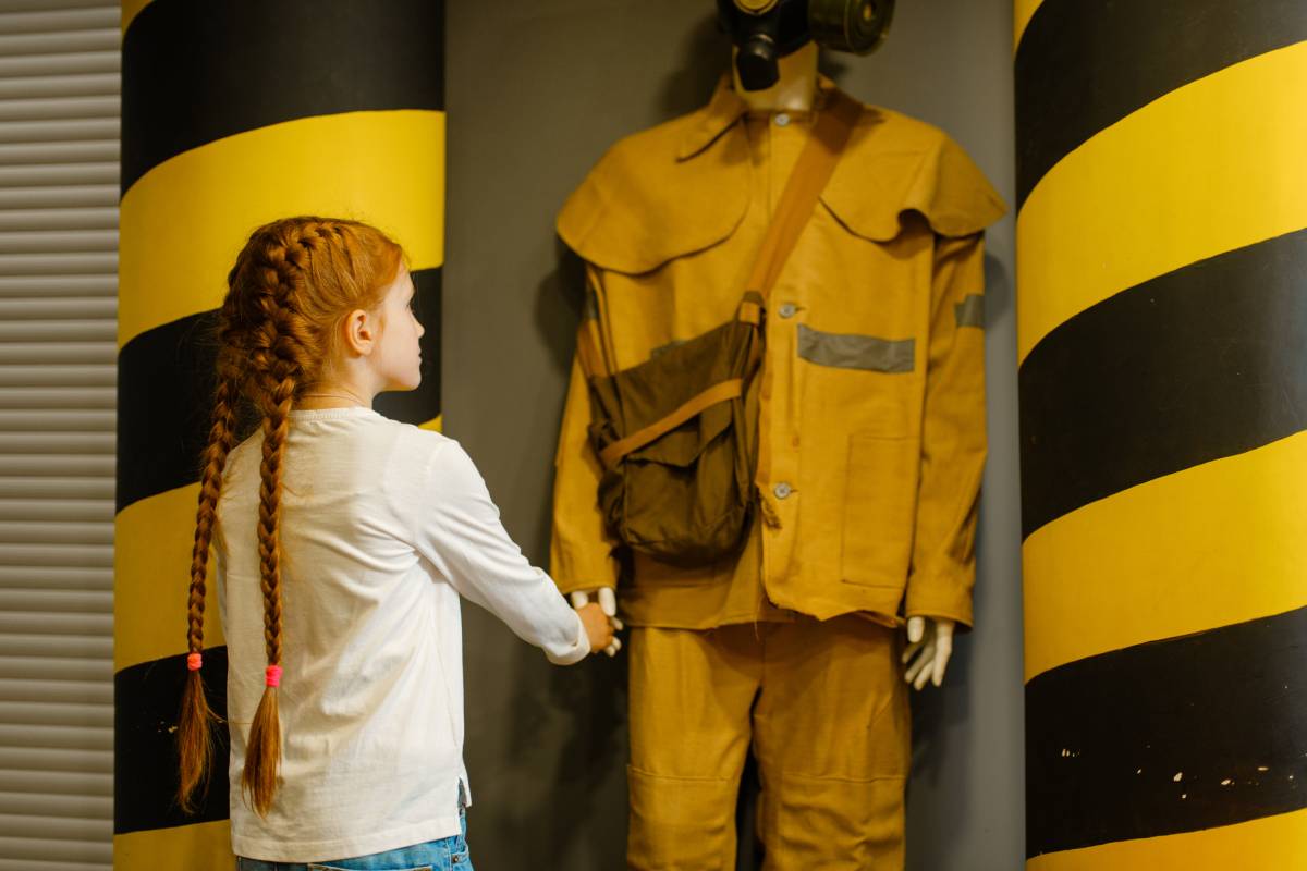 Female child looks on fireman mannequin in helmet and uniform, playroom indoor. Kid lerning firefighter profession. Lifeguard is a chilish dream