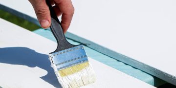 Male builder paints white boards for construction. Male hand holds brush. Painting work, house renovation.