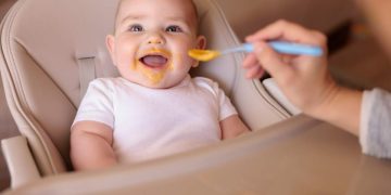 Mother feeding her cheerful baby boy while introducing first solid food meal, baby all messy and staied with porridge sitting in high chair and laughing