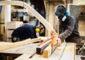 Man wearing ear protectors, protective goggles and dust mask standing in a warehouse, cutting piece of wood with circular saw.