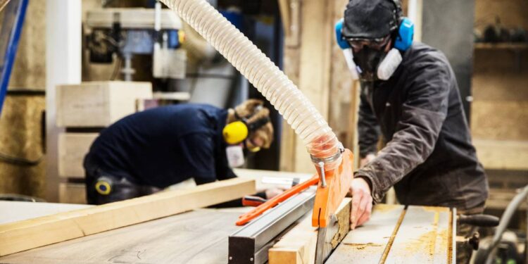 Man wearing ear protectors, protective goggles and dust mask standing in a warehouse, cutting piece of wood with circular saw.